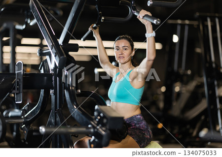 In gym, woman uses gym apparatus to strengthen back and shoulder muscles In gym, woman uses gym apparatus to strengthen back and shoulder muscles 134075033