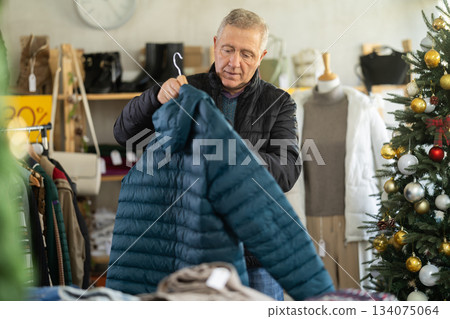 Elderly man chooses warm down jacket in clothing store. Elderly man chooses warm down jacket in clothing store. 134075064