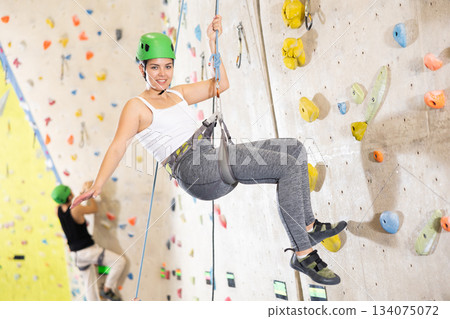 Sporty cheerful young female climber in helmet using safe rope going down after training in bouldering amusement center 134075072