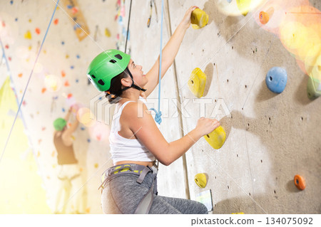 young girl with safety rope boldly climbs steep high artificial rock climbing wall in gym young girl with safety rope boldly climbs steep high artificial rock climbing wall in gym 134075092