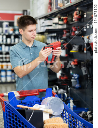 Man examines tool and buys electric drill, collects necessary supplies in store according to list for apartment repairs Man examines tool and buys electric drill, collects necessary supplies in store according to list for apartment repairs 134075102