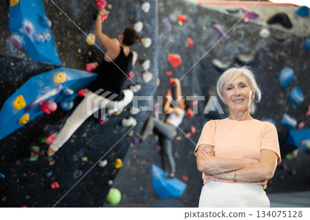 Elderly woman in sportswear smiling at camera while standing against artificial training climbing rock wall in adventure park 134075128