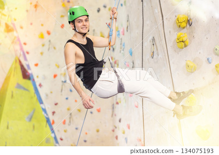 Well-equipped, positive young man going down on rope after reaching top in indoor bouldering adventure park 134075193
