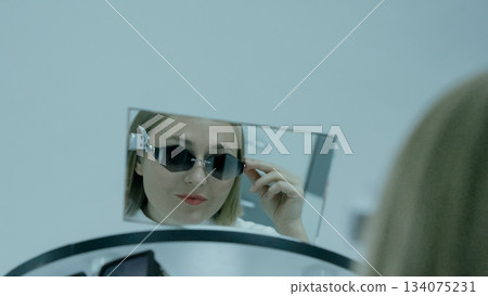 Young woman in glasses looks at her reflection in a large mirror at the store. Young woman in glasses looks at her reflection in a large mirror at the store. 134075231