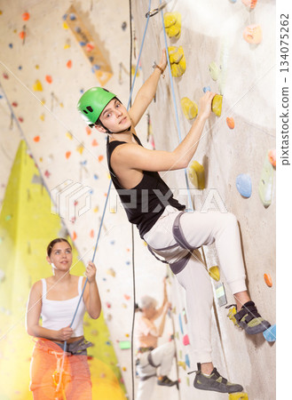 Well-equipped, positive young man training on stone wall, artificial mountain with rocks, climbing up in bouldering park indoors 134075262