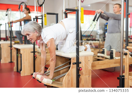 Senior woman does gymnastics in gym and performs bends and stretches on wunda chair Senior woman does gymnastics in gym and performs bends and stretches on wunda chair 134075335