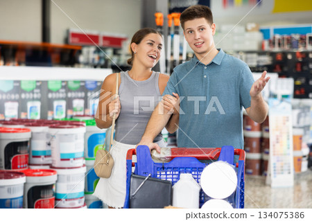 Positive married couple, young adult man and woman, shopping for materials and tools for house renovation, walking with trolley cart along aisles in hardware store 134075386