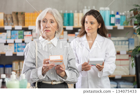 Senior woman choose antiallergic pills in pharmacy, woman pharmacist in background Senior woman choose antiallergic pills in pharmacy, woman pharmacist in background 134075433