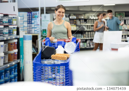 Girl with trolley is standing in sales area of paintwork material department of supermarket recalls position of goods needed for purchase. Male and female buyers in background Girl with trolley is standing in sales area of paintwork material department of supermarket recalls position of goods needed for purchase. Male and female buyers in background 134075439
