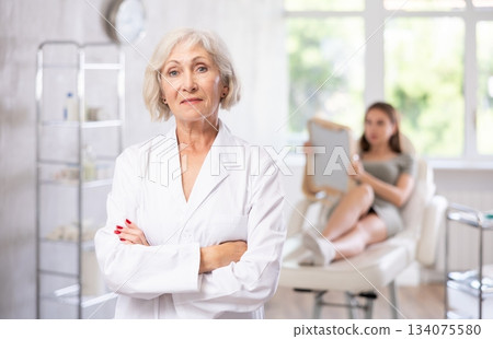Smiling senior woman doctor in throws up his hands with woman patient looking at mirror behind in clinic. 134075580