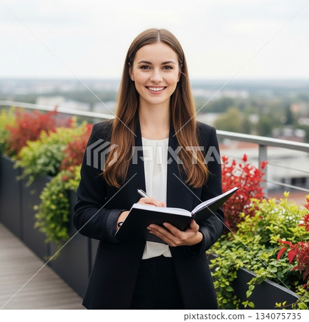 一位年輕的女商人面帶微笑，在戶外露臺上用筆記本寫字。 134075735