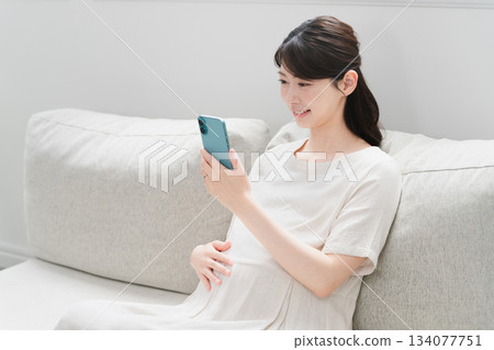 Pregnant woman operating a smartphone on the sofa Lifestyle image Pregnant woman operating a smartphone on the sofa Lifestyle image 134077751