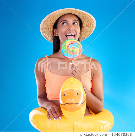 Pool float, happy woman and lollipop lick in a studio with sweet, swimsuit and sweets. Isolated, blue background and holiday of a young female with happiness, licking candy and feeling funny or silly 134078398