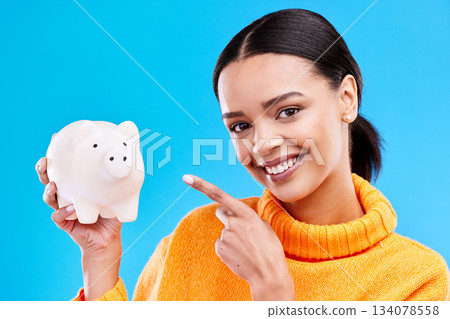 Happy woman, money and pointing to piggy bank for investment, budget or finance against blue studio background. Portrait of female with savings, cash or coin for profit investing or financial freedom 134078558