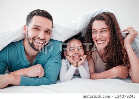 Family in bed, happy parents and child in portrait, relax and smile together with love and care. Spending quality time under blanket, bonding and relationship, man and woman with young girl at home 134079243