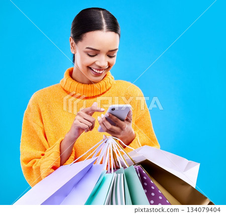 Happy woman, phone and shopping bags with smile for purchase, sale or discount against a blue studio background. Excited female shopper smiling on mobile smartphone holding gift bag for social media 134079404