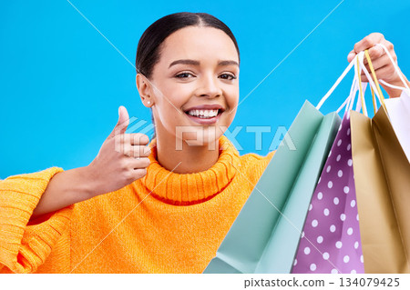 Happy woman, face and shopping bags with thumbs up for purchase, sale or discount against a blue studio background. Portrait of female shopper holding gift bag showing thumb emoji, yes sign or like Happy woman, face and shopping bags with thumbs up for purchase, sale or discount against a blue studio background. Portrait of female shopper holding gift bag showing thumb emoji, yes sign or like 134079425