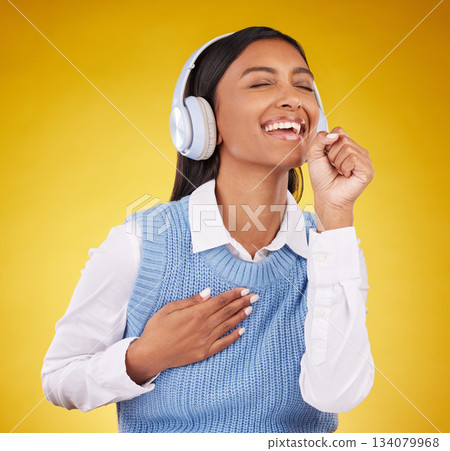 Headphones, happy and woman singing in studio while listening to music, playlist or album. Happiness, smile and Indian female model doing karaoke while streaming a song isolated by yellow background. 134079968