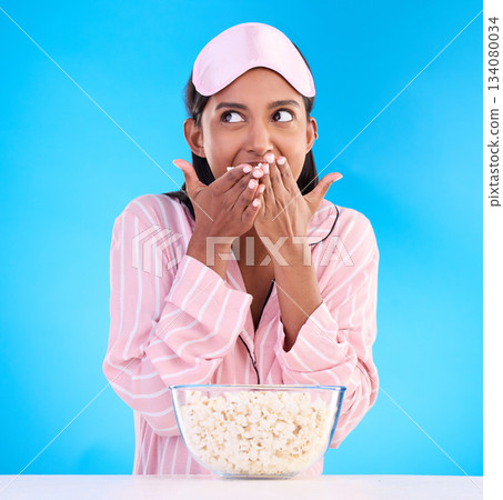 Popcorn, pajamas and thinking with a woman on a blue background in studio watching a movie for entertainment. Idea, video and night with an attractive young female eating a snack while streaming Popcorn, pajamas and thinking with a woman on a blue background in studio watching a movie for entertainment. Idea, video and night with an attractive young female eating a snack while streaming 134080034