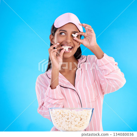 Popcorn, pajamas and playful with a woman on a blue background in studio watching a movie for entertainment. Fun, video and night with an attractive young female eating a snack while streaming Popcorn, pajamas and playful with a woman on a blue background in studio watching a movie for entertainment. Fun, video and night with an attractive young female eating a snack while streaming 134080036
