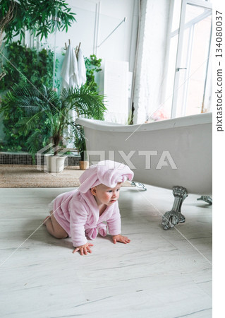 Moscow, Russia, 29.06.23. A baby in a pink robe is sitting on the floor in front of a bathtub Moscow, Russia, 29.06.23. A baby in a pink robe is sitting on the floor in front of a bathtub 134080037