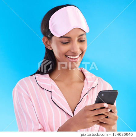 Happy, bedtime and a woman typing on a phone isolated on a blue background in studio. Smile, reading and an Indian girl with social media notification, mobile chat or online communication in pyjamas Happy, bedtime and a woman typing on a phone isolated on a blue background in studio. Smile, reading and an Indian girl with social media notification, mobile chat or online communication in pyjamas 134080040