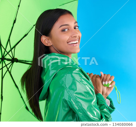 Happy, portrait and woman with an umbrella in studio with a stylish, trendy and fashion green rain coat. Life insurance, smile and Indian female model with a winter outfit isolated by blue background Happy, portrait and woman with an umbrella in studio with a stylish, trendy and fashion green rain coat. Life insurance, smile and Indian female model with a winter outfit isolated by blue background 134080066
