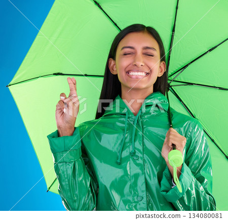Green, umbrella and fingers crossed with a woman in studio on a blue background during winter for insurance. Rain, fashion or cover and an attractive young female wishing for weather change with hope 134080081