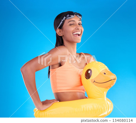 Portrait, wink and rubber duck with a woman on a blue background in studio excited for summer. Happy, smile and playful with an attractive young female ready for swimming on holiday or vacation 134080114