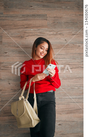 Phone, fashion and a woman in studio on a wooden background typing a text message or reply. Mobile, contact and attractive young female browsing social media while carrying a handbag in red clothes Phone, fashion and a woman in studio on a wooden background typing a text message or reply. Mobile, contact and attractive young female browsing social media while carrying a handbag in red clothes 134080676