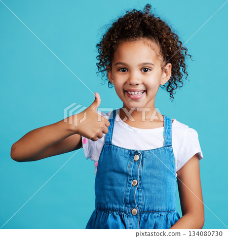 Happy, portrait and girl child with thumbs up in a studio with success, happiness and achievement. Smile, positive and face of a kid with a satisfaction or approval hand gesture by a blue background. Happy, portrait and girl child with thumbs up in a studio with success, happiness and achievement. Smile, positive and face of a kid with a satisfaction or approval hand gesture by a blue background. 134080730
