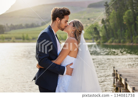 Love, wedding and a married couple kissing by a lake outdoor in celebration of their marriage for romance. Water, summer or kiss with a bride and groom bonding together in tradition after ceremony 134081738