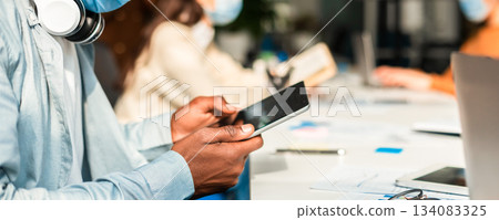 Education And Work During Pandemic. Closeup of african american man wearing surgical face mask and using digital tablet, sitting at desk. Group of diverse people sitting at table in blurred background Education And Work During Pandemic. Closeup of african american man wearing surgical face mask and using digital tablet, sitting at desk. Group of diverse people sitting at table in blurred background 134083325