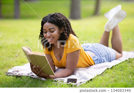 Happy African American woman reading interesting book while lying on green lawn at park Happy African American woman reading interesting book while lying on green lawn at park 134083340