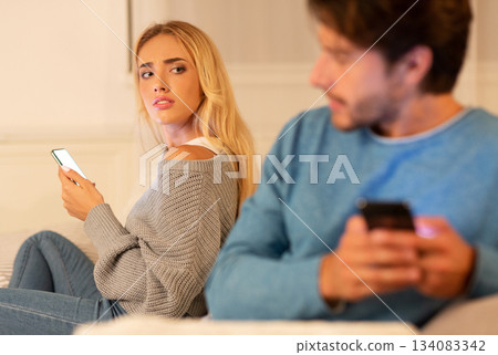 Cheating And Infidelity. Husband Texting On Cellphone Sitting Next To Suspicious Wife On Couch At Home. Selective Focus Cheating And Infidelity. Husband Texting On Cellphone Sitting Next To Suspicious Wife On Couch At Home. Selective Focus 134083342