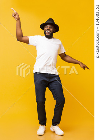 Cheerful black man in hat dancing and pointing index fingers aside on yellow studio background 134083353