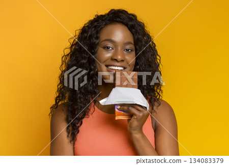 Attractive african girl holding milk chocolate next to her lips and smiling, yellow background Attractive african girl holding milk chocolate next to her lips and smiling, yellow background 134083379