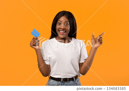Pretty young black lady with credit card pointing at copy space over orange studio background. Smiling african american woman showing empty space for advertisement, online shopping concept 134083385