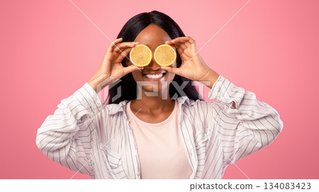 Happy young black lady with lemon halves in front of her eyes posing on pink studio background, panorama. Positive millennial woman with citrus fruits. Natural vitamins for skin detox concept 134083423