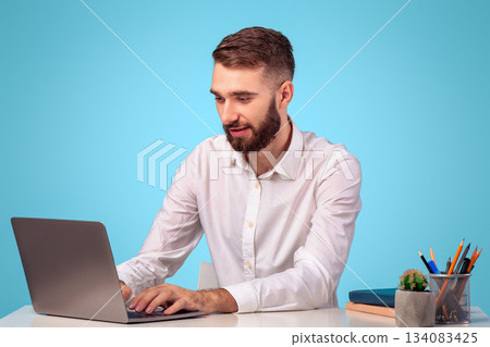 Online job concept. Attractive young businessman using laptop computer at his desk on blue studio background. Focused millennial office employee working on pc, typing, browsing internet 134083425