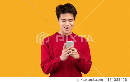 People And Technology Concept. Portrait of happy smiling young asian man using smartphone standing isolated over orange studio background, copy space. Guy chatting online, browsing social media 134083428