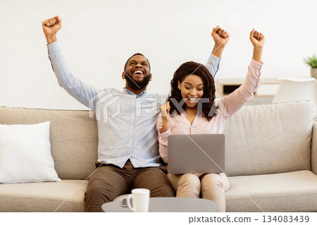 Lucky Winners. Excited enthusiastic african american couple using laptop computer, celebrating online win and shaking fists sitting on the sofa at home. Happy man and woman reading great news 134083439