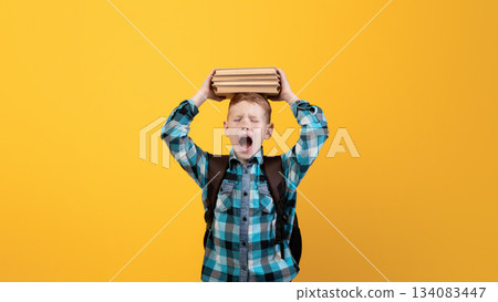Stressed schoolboy with stack of books on his head yelling on yellow studio background, got tired of studying, need for rest, copy space. Ginger boy holding books on his head and screaming 134083447