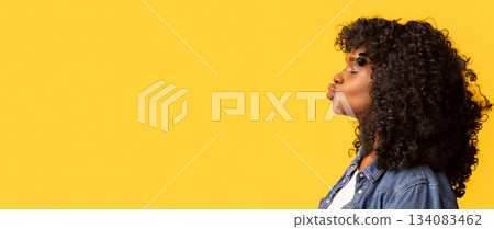 Side view of emotional beautiful african american young lady with booshie hair showing kissing lips towards copy space over yellow studio background, panorama. Love, emotions, feelings, kissing face 134083462