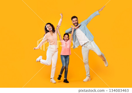 A family of three is posing for a photo in a studio. The father is striking a playful pose with his arm outstretched, while the mother and daughter are jumping and smiling brightly. 134083463