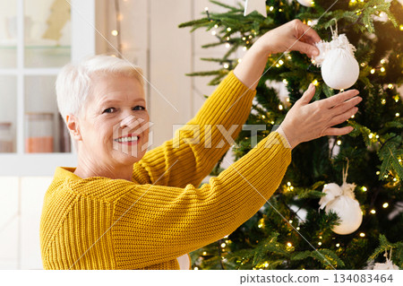 Smiling elderly woman decorating Christmas tree. kitchen interior, copy space. Beautiful old lady with short haircut making Christmas decorations before winter holidays, getting ready for family party 134083464