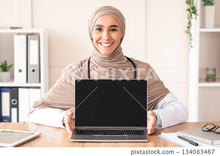 Happy female doctor muslim woman in hijab showing laptop with blank screen, mockup, copy space. Smiling doctor using modern technologies in her job, working and training online, clinic interior 134083467