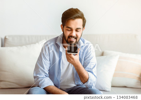 Smiling Arab Man Recording Voice Message On Smartphone Or Using Virtual Assistant While Relaxing On Couch At Home, Eastern Millennial Guy Enjoying Smart Technologies For Communication, Copy Space 134083468