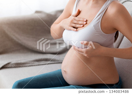 Unrecognizable pregnant lady massaging her breasts while sitting on couch at home, expectant female having painful feelings in chest area, examinating herself, cropped image with free space Unrecognizable pregnant lady massaging her breasts while sitting on couch at home, expectant female having painful feelings in chest area, examinating herself, cropped image with free space 134083486