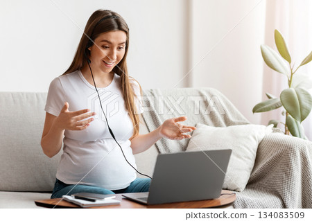 Web Conference. Cheerful pregnant woman in headset having video call on laptop at home, talking and gesturing at camera while sitting on couch, happy expectant lady enjoying online communication 134083509